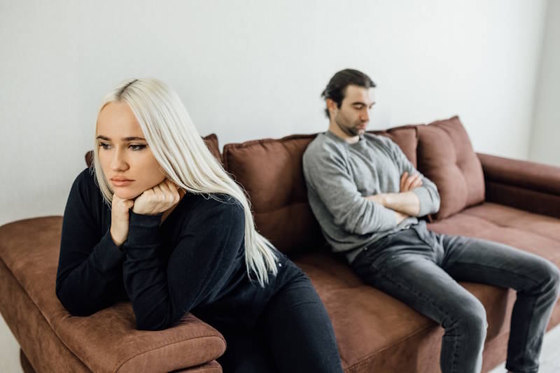 Couple assis sur un canapé, tourné l’un contre l’autre, exprimant la distance émotionnelle et la méfiance qui naissent de la jalousie dans le couple.
